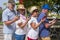 Four senior people looking at each his cell phone standing under palm trees and flowering plants in a public park - relaxed and