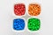 Four squared bowls with small red, yellow, blue, green and orange coated chocolate candies similar to m&ms in a squared bowl isola