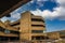 A four story brown concrete office buildings with tall ball shaped light posts and blue sky with powerful clouds