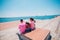 Four tourist friends having fun and taking pictures on a modern abstract bench paced on a seaside marine port on a sunny day.