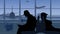 The frame shows an airport with a waiting room. Two people in silhouette are sitting on opposite sides, a man is taking