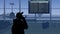 The frame shows an airport with a waiting room. A woman in silhouette looks at the scoreboard and checks the flights