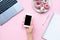 Frame woman`s hands holding white smartphone with black copyspace on pink background with other accessories. Feminine business mo
