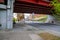 A freeway underpass with homeless people sleeping underneath with man walking along the sidewalk with lush green trees