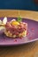 Fresh beef steak tartare with parmesan cheese, capers, croutons and yolk quail on a purple plate on a wooden background