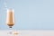 Fresh coffee beverage with cream, biscuits, straw in wineglass in blue interior on white wooden table with copy space.