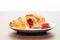 Fresh french croissant with strawberry jam filling cut into two pieces and strawberry on white ceramic plate on bright light brown