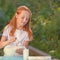 Fresh milk pours from a jug into a glass a red-haired girl. Warm sunlight illuminates the girl and the milk on the table. Glass of