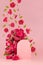 Fresh pink roses as arch, empty rounded door as podium with green leaves, buds fly as swirl on abstract pink scene mockup.