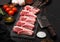 Fresh raw butchers lamb beef cutlets on stone board with vintage meat fork and knife and hatchet on black background.Salt, pepper