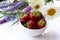 Fresh strawberries in a bowl with bouquet of summer flowers on white table. Top view, copy space.