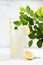 Fresh summer classic lemonade in glass with straw, ice, lemon slice in soft light modern white kitchen interior with marble tile.