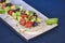 Fresh vegetables and green salad with sauce served on traditional rectangular white plate over bright blue background.