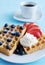 Fresh Viennese or Belgian waffles on a porcelain plate with fresh strawberries and blueberries and whipped cream and a cup of