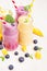 Freshly blended yellow and violet fruit smoothie in glass jars with straw, mint leaves, mango slices, blueberry, close up. Soft w