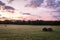 Freshly rolled hay bales rest on rolling hill with dramatic cloudscape at sunrise