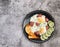 Fried eggs with green onions, sausages, toast and cucumber slices on a round plate on a dark grey background