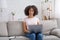 Friendly african american female working in laptop, sitting on couch with notepad in living room interior