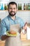 Friendly bearded waiter smiling while pointing to the tasty sandwich