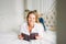 Friendly charming blonde woman with long fair hair in casual clothing reading book on bed in bright rich interior