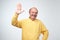 Friendly-looking attractive european pensioner in yellow t-shirt waives hand in hello gesture while smiling cheerfully.