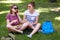 Friendly looking positive female fellows speak with each other, gesture with hands, keep legs crossed, rest on green grass, enjoy