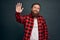 Friendly modest handsome bearded man in red checkered shirt saying hi with hesitation lifting one palm, waving hand in hello,