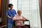 Friendly staff caregiver of nursing home talking to asian senior man in living room