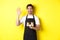 Friendly waiter in cafe waving hand at customer, holding takeaway coffee oder, standing against yellow background in