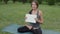 Friendly woman yoga trainer with blank sheet in hands looking at camera while sitting cross-legged on mat in park.