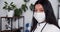 Friendly young multiethnic doctor woman wearing mask, white coat smiling at camera at light modern clinic workplace.