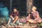 Friends camping in forest. Two playful girls in vintage dresses looking at lumberjack. Guy with serious look sitting by