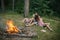 Friends relax at campfire. People at bonfire flame in green forest. Women and man at fire in camp. Camping, hiking and