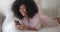 A frizzy-haired, good-looking afro american woman lies on a bed with a smartphone