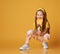 Frolic active red-haired kid girl in yellow t-shirt and colorful leggings sits squatting, eating, biting big lollipop