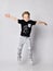 Frolic blond kid boy in sunglasses, black t-shirt with dinosaur print and gray pants jumps with hands spread wide