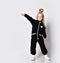 Frolic blonde kid baby girl with braids in stylish black jumpsuit and sneakers stands and points at upper corner