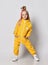 Frolic blonde kid girl with modern hairstyle in yellow jumpsuit and sneakers stands posing with her hands in pockets