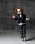 Frolic excited kid girl in leather jacket, jeans and sunglasses is having fun jumping singing like a rock star on gray