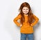 Frolic red-haired kid girl in orange sweatshirt and blue jeans stands with fists at waist playing displeased strict mom