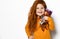 Frolic smiling five-year-old red-haired kid girl in orange sweatshirt holds small redhair doll in hands on her shoulder