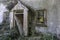 Front of derelict cottage with entrance door leaning to one side in small porch. With a broken window on either side. Vegetation o