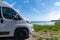 Front end of a gray camper van parked next to a picturesque shore with turquoise water and forest under a blue sky