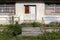 Front entrance to an abandoned house with destroyed windows and locked doors surrounded with plants covering concrete steps and