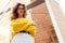 Front portrait of a girl with wavy hair, wear in yellow sweater and white jeans behind modern building with cross arm.