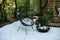 Front veranda of house with two black Acapulco armchairs, coffee table and plants pots. Metal black fireplace bowl in garden on ba