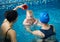 Front view of baby boy during swimming lesson. Back view of mother holding child and woman pouring water on child`s head
