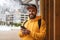 Front view. Bearded male tourist in yellow hoodie and cap with black backpack stands outdoors and using smartphone. Lifestyle