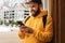 Front view. Bearded male tourist in yellow hoodie and cap with black backpack stands outdoors and using smartphone. Lifestyle