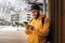 Front view. Bearded male tourist in yellow hoodie and cap with black backpack stands outdoors and using smartphone. Lifestyle